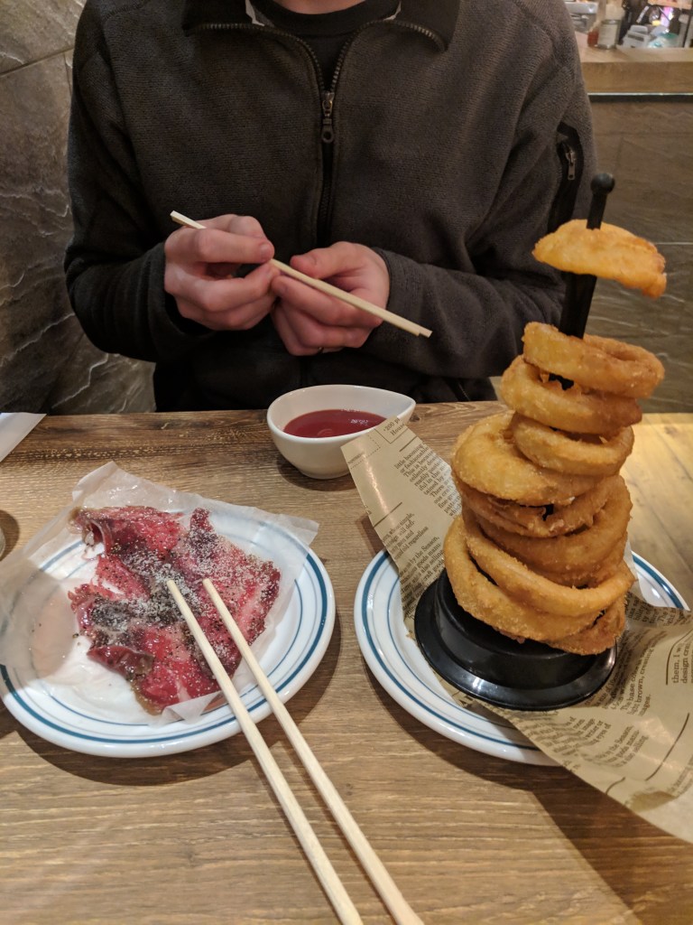 A person holding chopsticks above a plate of raw meat and a small bowl of dipping sauce, with a stack of onion rings on a stand nearby.