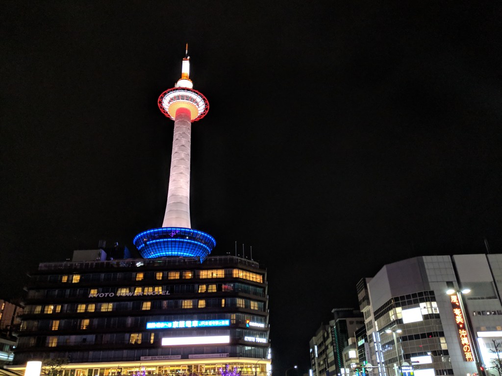 Kyoto Tower illuminated at night, featuring red, white, and blue lights against a dark sky.