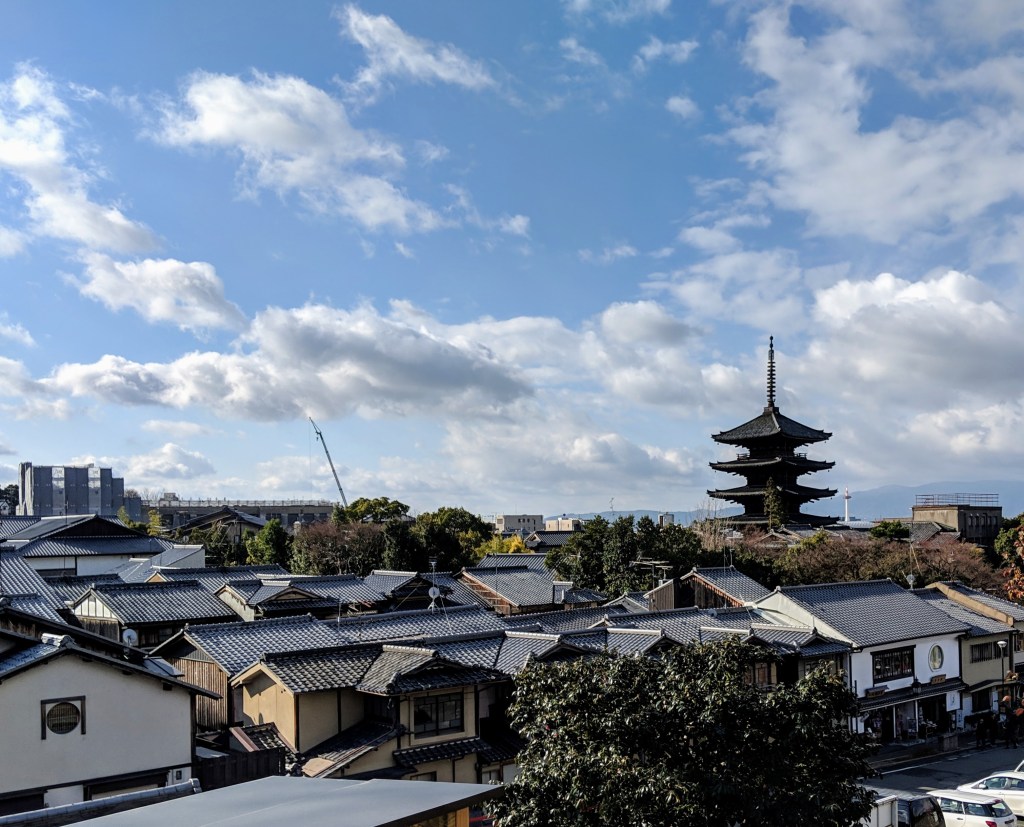 A panoramic view of traditional Japanese architecture with rooftops, featuring a pagoda in the background against a blue sky with fluffy clouds.