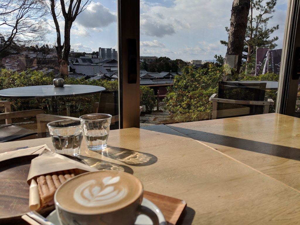 A cozy café scene featuring a latte with a leaf design, a plate of cookies, and two glasses of water on a wooden table, with a view of trees and rooftops through the large window.