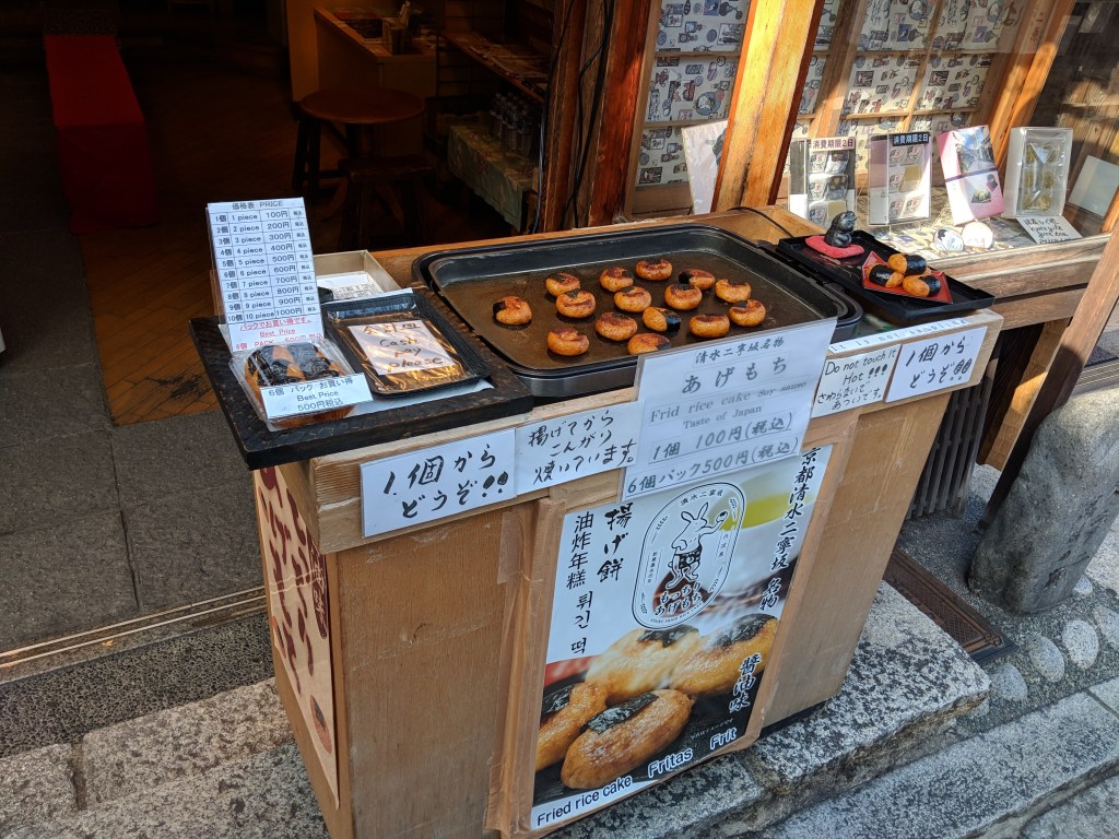 Street vendor display selling fried rice cakes in Japan, featuring a tray of round fried items. Price list for individual and multiple pieces is visible, along with a sign inviting customers to try the treats.