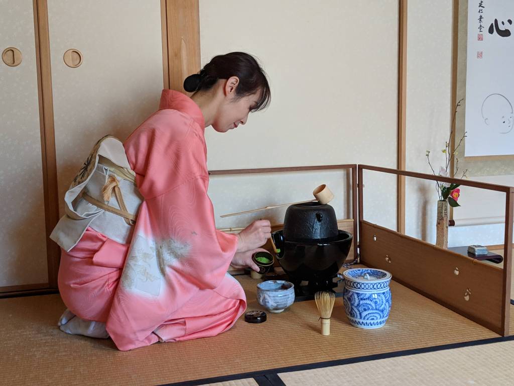 A woman in a pink kimono preparing tea in a traditional Japanese tea ceremony setting, kneeling beside a black tea pot and surrounded by utensils on a tatami mat.