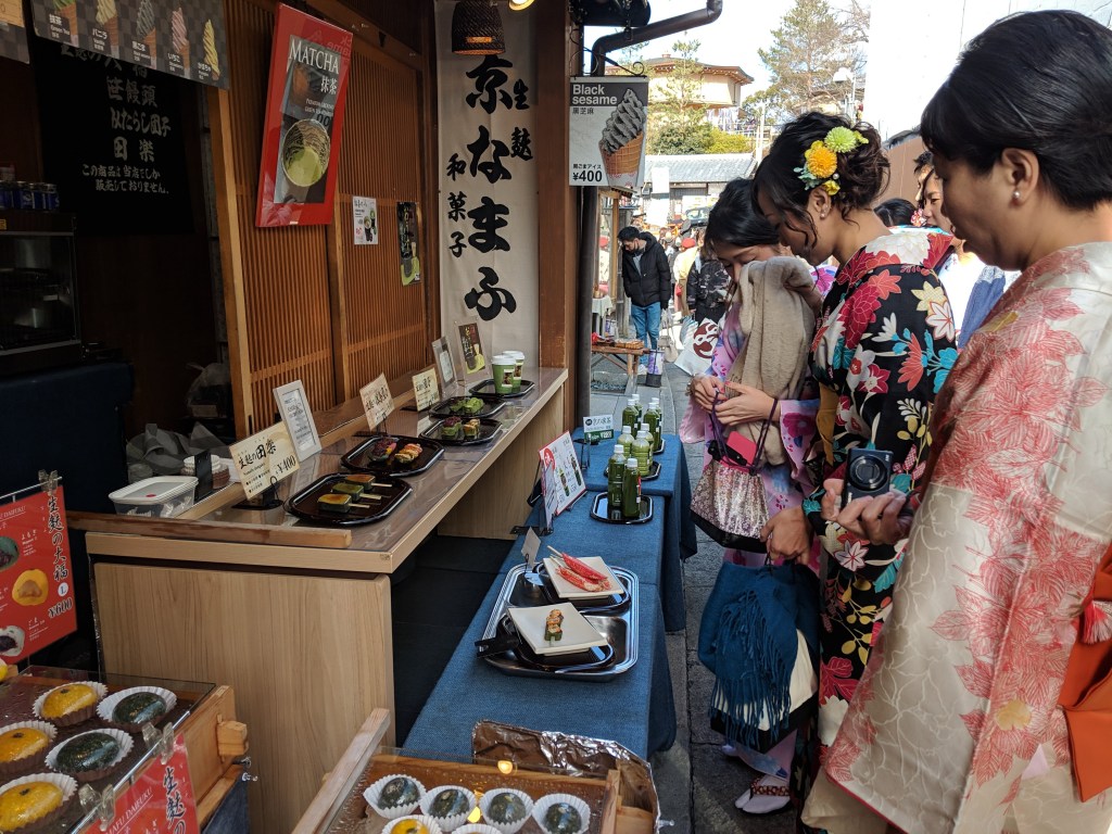 A traditional food stall in Japan featuring various dishes like matcha and sushi, with customers dressed in kimono examining the offerings.