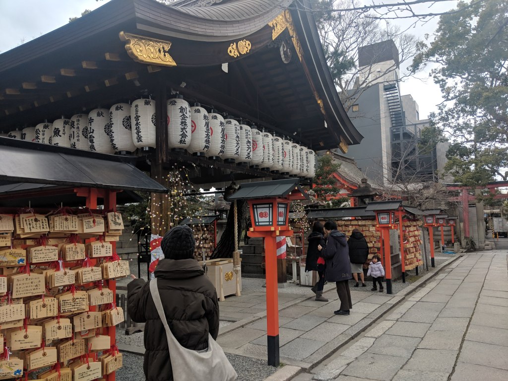 A traditional shrine entrance adorned with hanging lanterns, surrounded by visitors. Wooden plaques with wishes are displayed on stands nearby, creating a serene atmosphere in an urban setting.