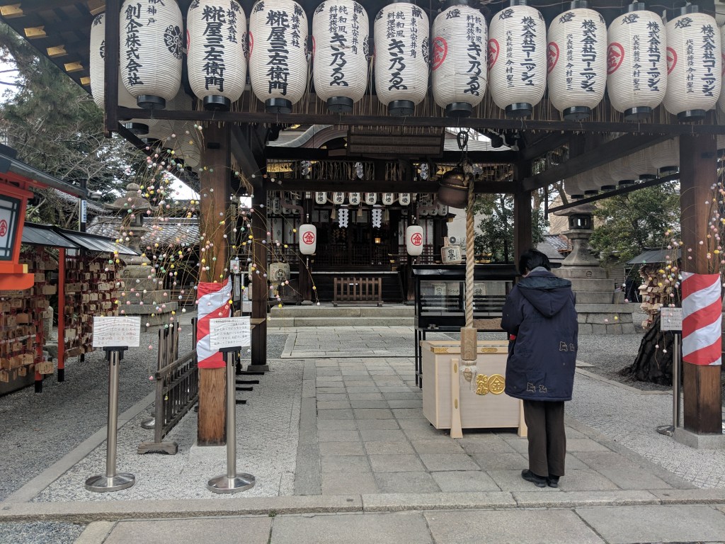 A person standing in front of a traditional Japanese shrine, with decorative lanterns hanging above and a wooden offering box visible nearby.