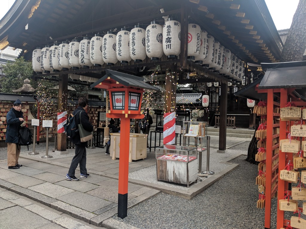 A traditional Japanese shrine entrance featuring hanging paper lanterns, wooden prayer plaques, and a decorative lantern post, with visitors interacting in the vicinity.