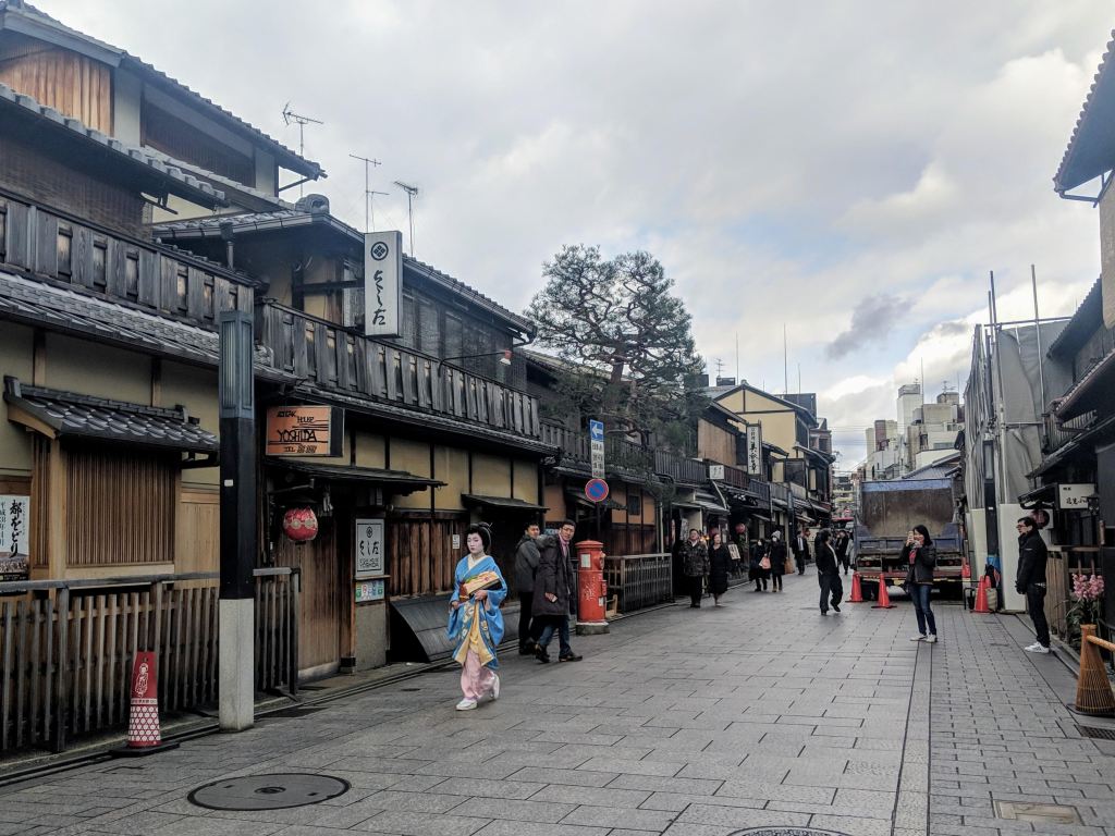 Traditional street in a historic area of Japan, featuring wooden buildings, a person in a kimono, and pedestrians walking along a cobblestone pathway.