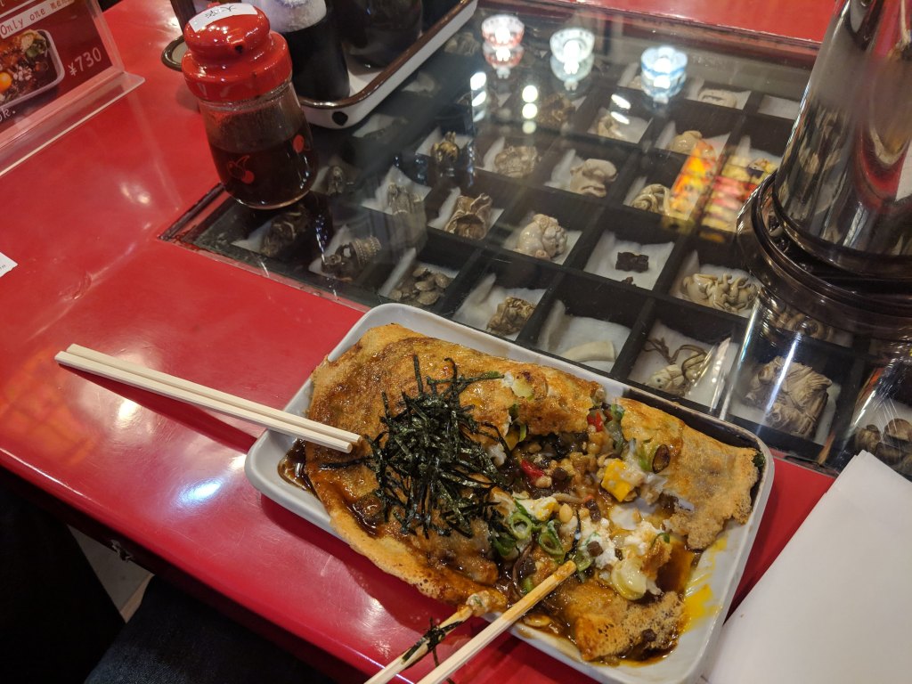 A plate of okonomiyaki topped with seaweed and various ingredients, served with chopsticks on a red table.