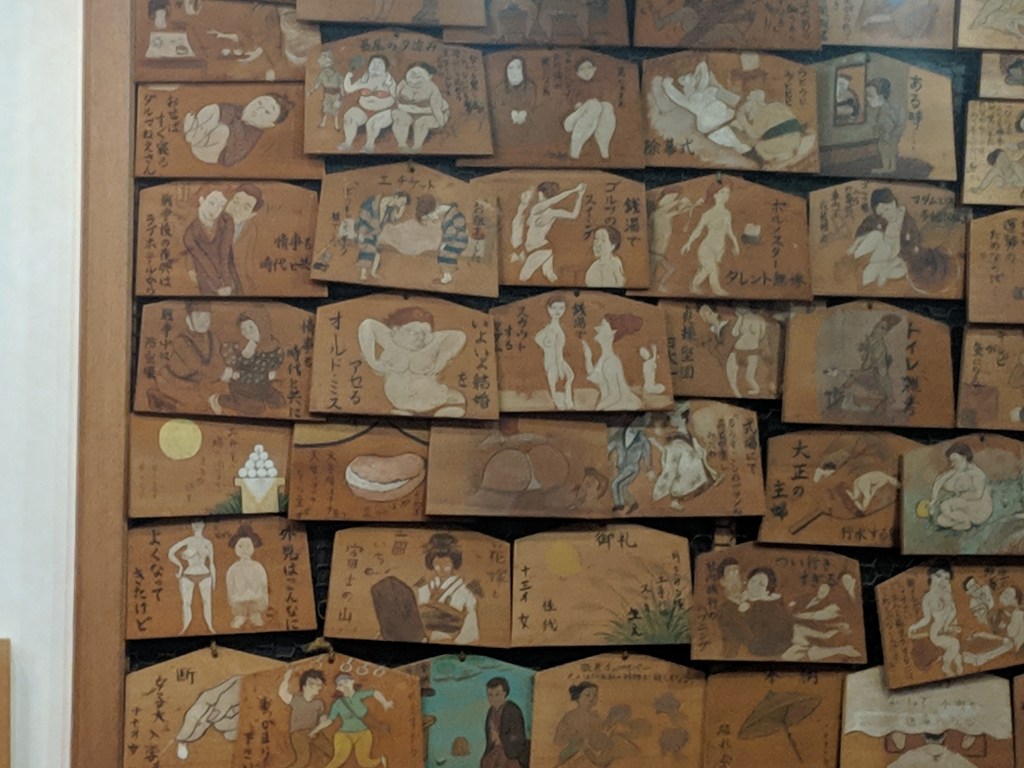 A collage of wooden plaques, each displaying illustrations of various scenes, including figures in playful or intimate poses, accompanied by Japanese text. The artwork features a mix of humor and cultural commentary.
