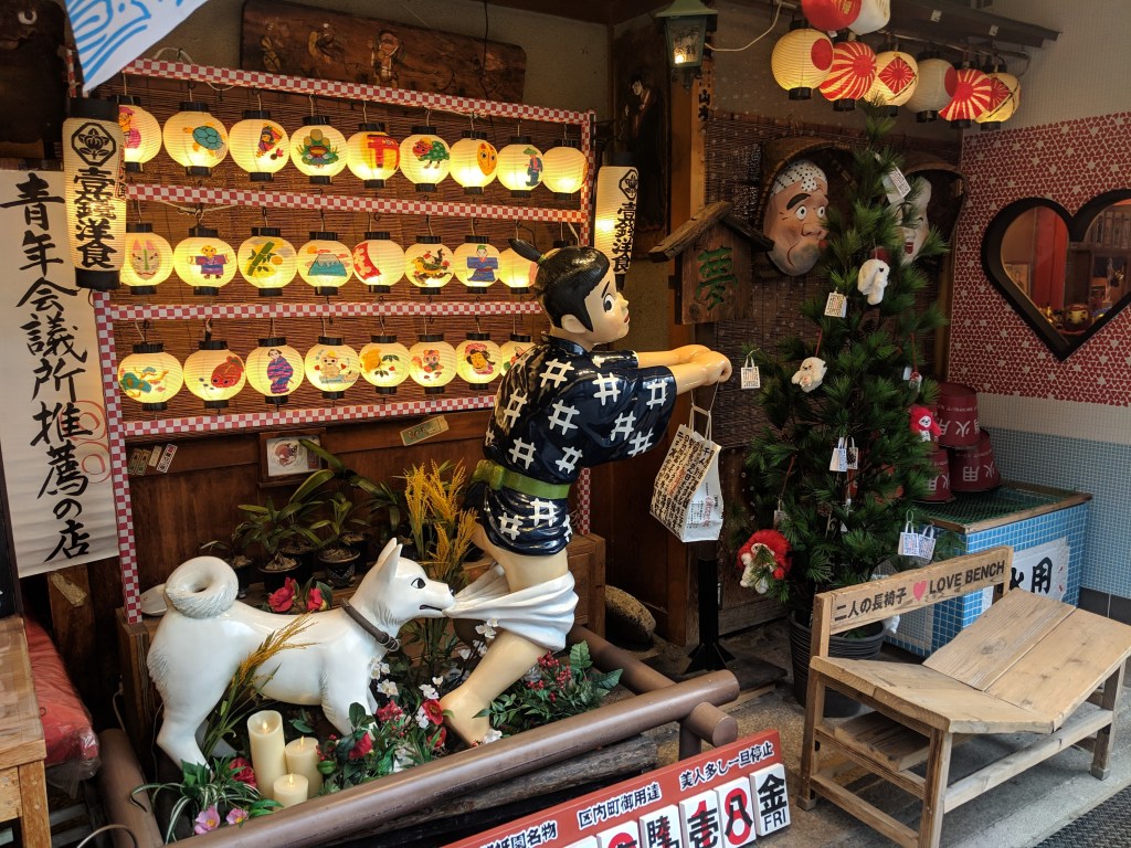 A traditional Japanese street scene featuring decorative lanterns, a statue of a boy in a kimono with a dog, and a small Christmas tree. A wooden bench and floral decorations are also visible.