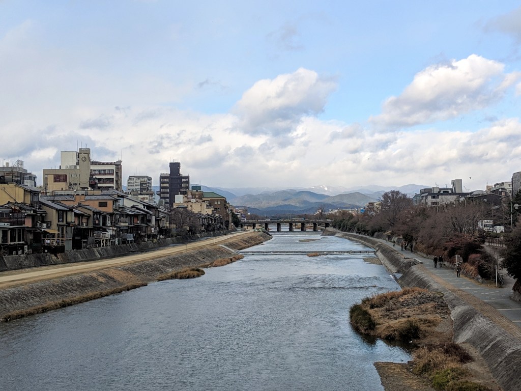 A scenic view of a river bordered by traditional and modern buildings, with mountains in the background under a partly cloudy sky.