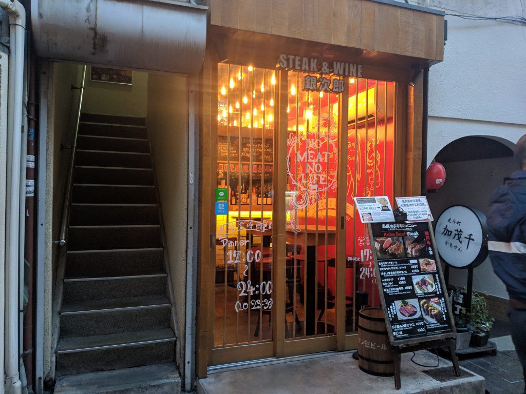 Entrance of a steak and wine restaurant with illuminated signs and a menu feature outside. A staircase is visible on the left.