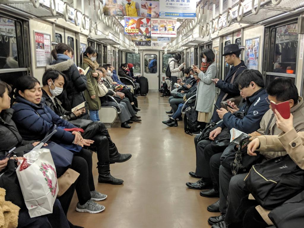 Interior view of a subway train with passengers seated and standing, some using mobile devices, others engaged in conversation, showcasing a typical commuting scene.