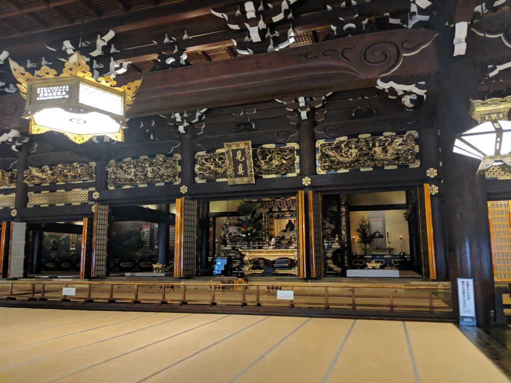 Interior view of a traditional temple featuring ornate wooden architecture, decorative carvings, a raised tatami mat floor, and a central altar with flowers and candles.