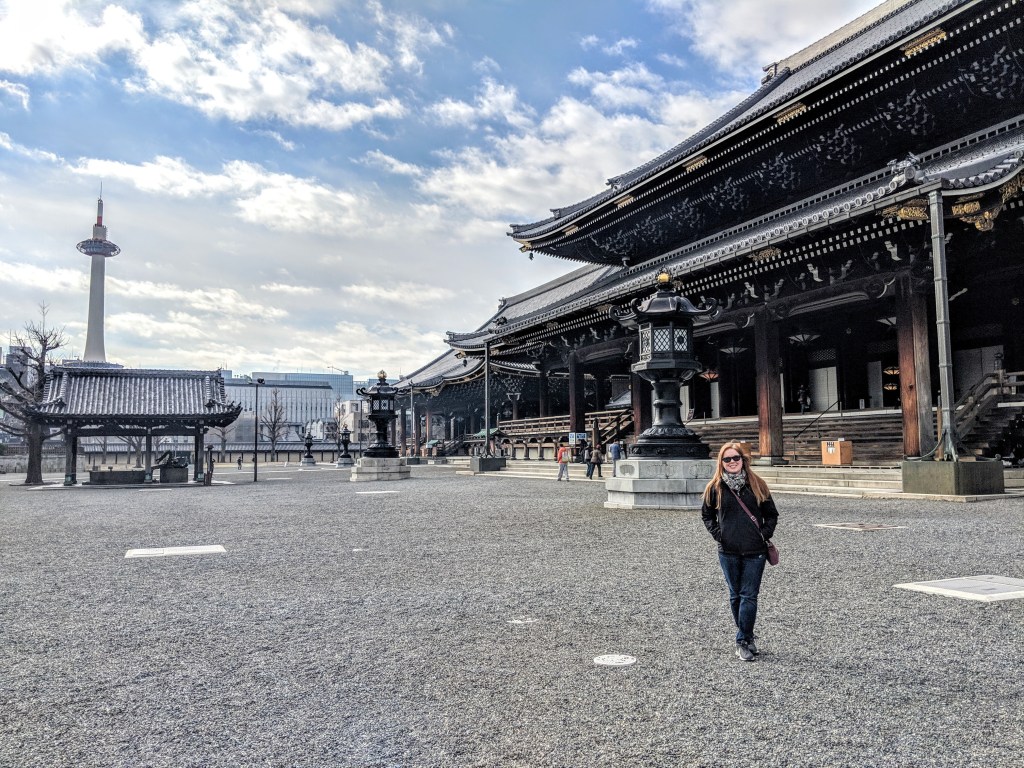 A person standing in a gravel courtyard in front of a traditional Japanese temple with ornate wooden architecture, and the Kyoto Tower visible in the background under a partly cloudy sky.