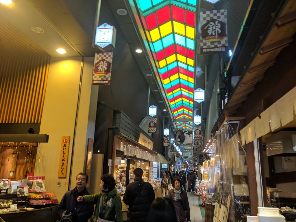 A bustling market street featuring various shops and stalls, with colorful stained glass overhead and people browsing.