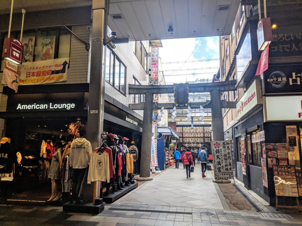 A narrow shopping street with Japanese architecture, featuring shops, clothing displays, and a traditional torii gate at the end.