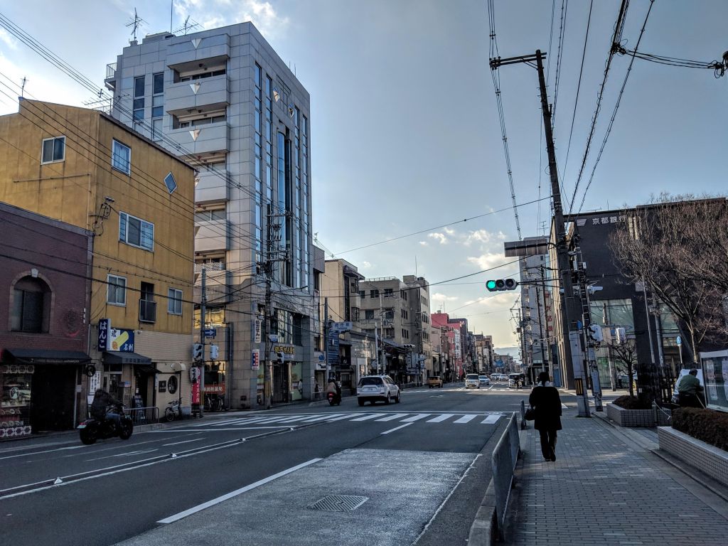 A city street view featuring a mix of modern and traditional buildings, with a pedestrian walking on the sidewalk and traffic lights in the background.