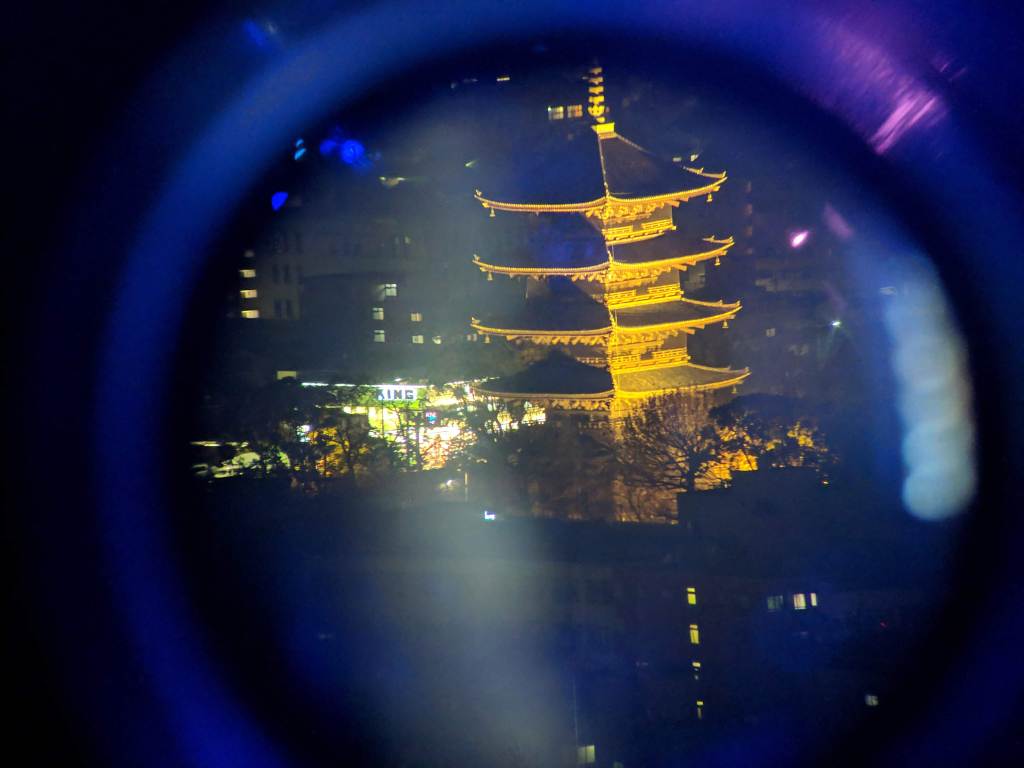 A night view of a lit-up pagoda seen through a circular lens, with surrounding buildings in the background.