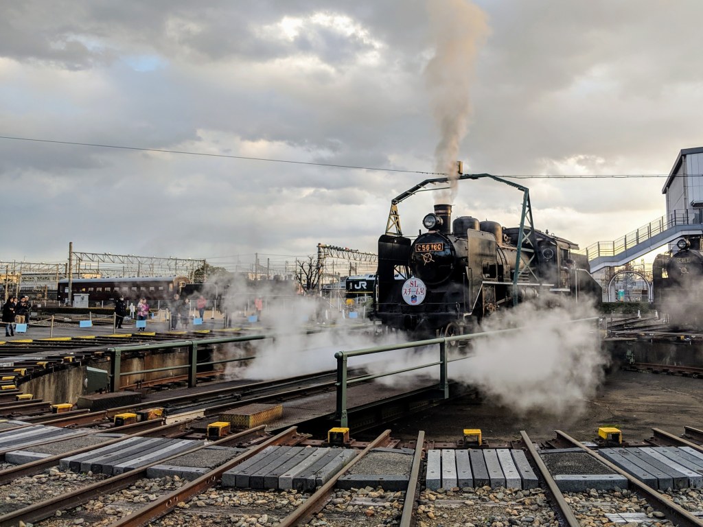 A vintage steam locomotive releasing steam at a train station, with spectators nearby and cloudy skies in the background.