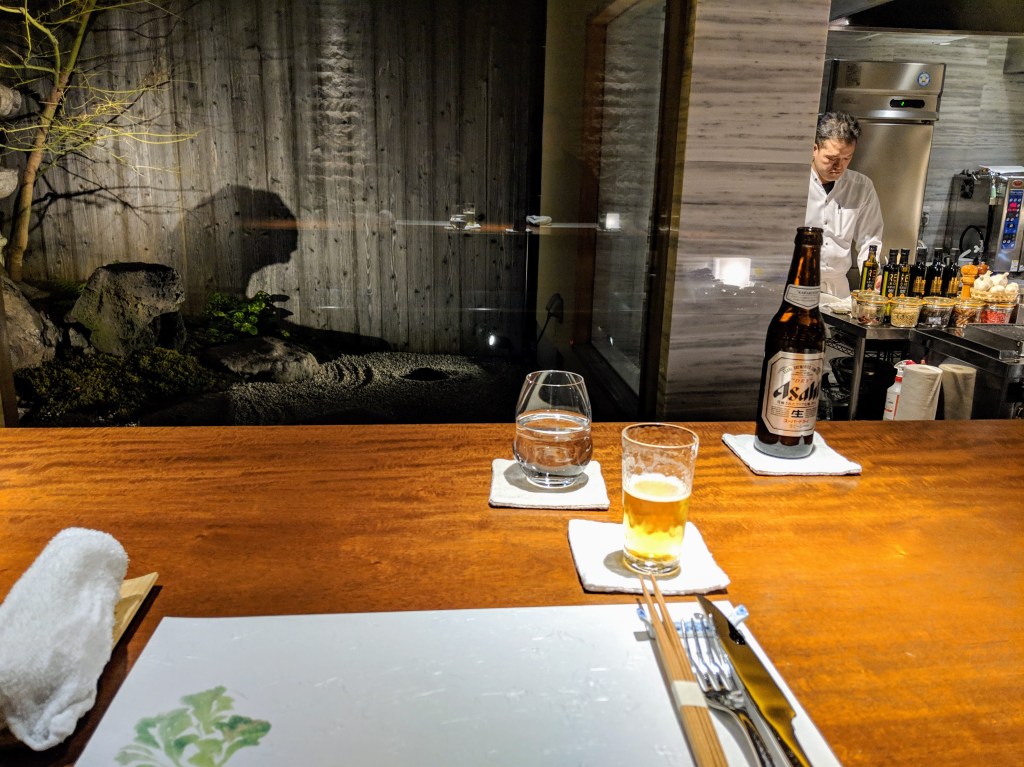 Wooden dining table setting with chopsticks and a plate, featuring a glass of water and a beer bottle. In the background, a kitchen area and a serene outdoor garden visible through a glass wall.