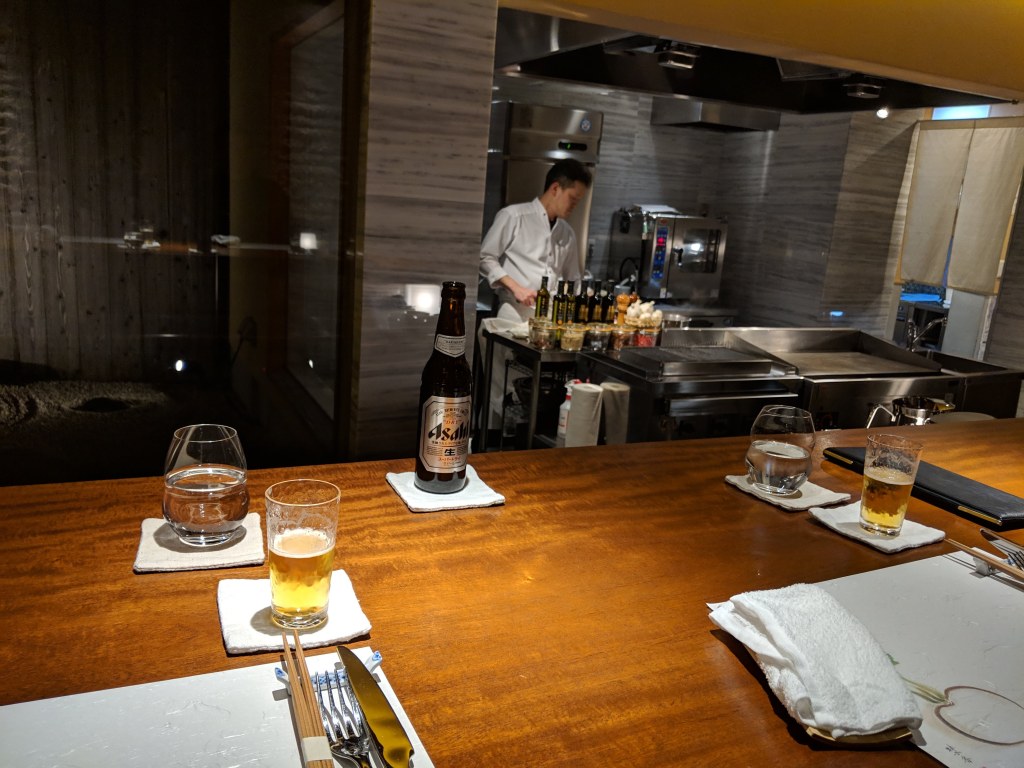 Counter view of a restaurant kitchen with a chef in the background, featuring a wooden table set with glasses, a bottle of beer, and utensils.