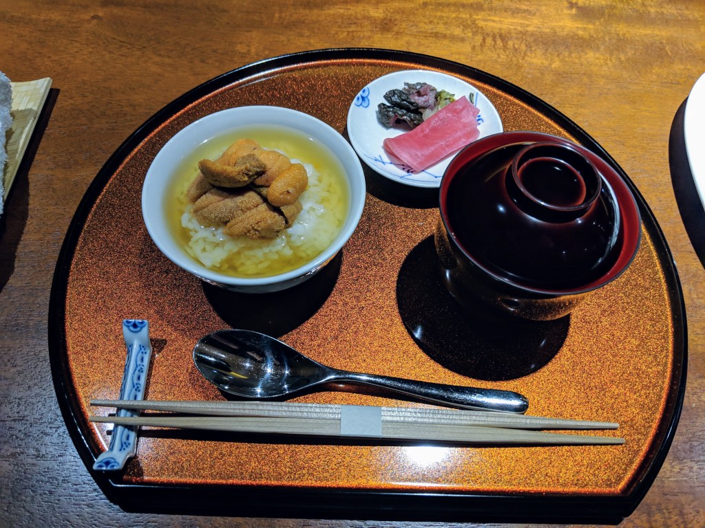 A bowl of rice topped with sea urchin served in a small dish on a decorative tray with a spoon and chopsticks, accompanied by a side plate of sliced tuna and pickled vegetables.