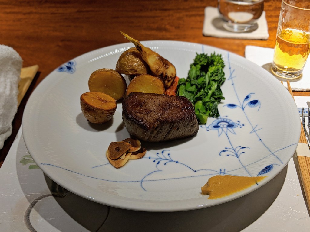 A plate featuring a portion of seared beef steak accompanied by roasted potatoes, colorful vegetables, and a dollop of sauce. A glass of drink and a napkin are visible in the background.